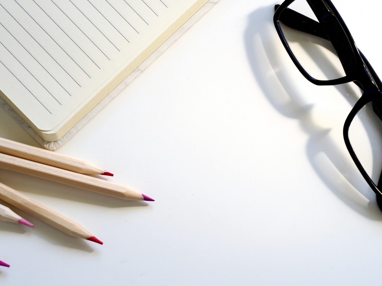 A clean desk setup showing a lined notebook, pencils, and black glasses on a white surface.
